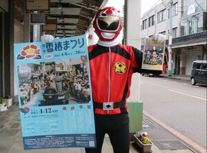 雪椿まつりのポスターを掲げ、なぞなぞラリーへの挑戦を呼びかける「小京都戦隊カモレンジャー」のレッド=加茂市仲町