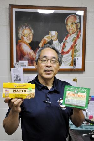 豆腐と納豆を手にする「アロハ豆腐ファクトリー」3代目社長のポール・ウエハラさん。祖父上原亀三郎さんと祖母鶴子さんを描いた絵が見守る=6月、ハワイ・ホノルル(共同)