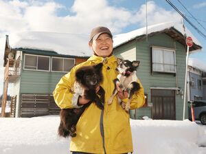 ２匹の愛犬を抱いて「津南・赤沢の家」の前に立つ宮沢悦子さん。「来訪者と地域をつなぎたい」と意気込む＝津南町赤沢