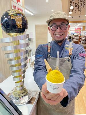 店舗前で世界一に輝いたマンゴーのジェラートを紹介する佐久間康之さん。隣は優勝トロフィー＝三条市福島新田