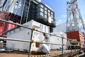 　福島第１原発１号機（左）前で作業する防護服を着た作業員。奥は２号機＝２月