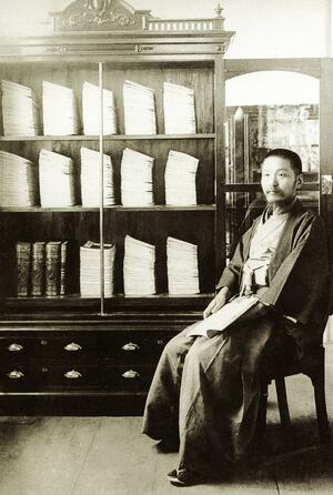 1907年完成の「大日本地名辞書」の原稿が積まれた書棚を前に、写真に収まる43歳の吉田東伍(吉田東伍記念博物館提供)