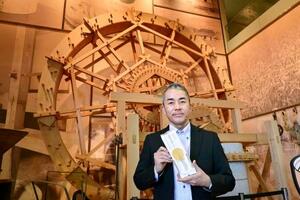 商品を持つ津村孝幸さん。「通常のうどんに比べてもっちりした食感の商品です」と話す。奥は戦前に使った製粉用の水車を復元した展示品=高松市
