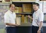 「大善寺国民学校」の日誌を手にする市川昌庸さん(左)と、日誌が見つかった書棚を示す福岡県久留米市立大善寺小の石橋敬嗣校長=8月6日、久留米市